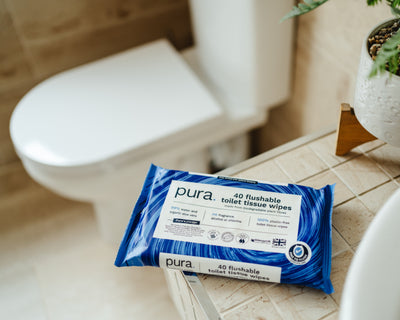 Pura flushable toilet tissue wipes package on a bathroom floor with a toilet and plant in the background.